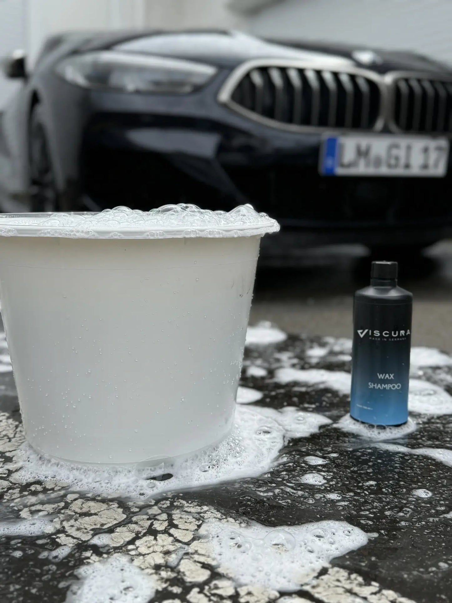 Viscura Wax Shampoo bottle next to a bubble-filled bucket, with a car in the background.