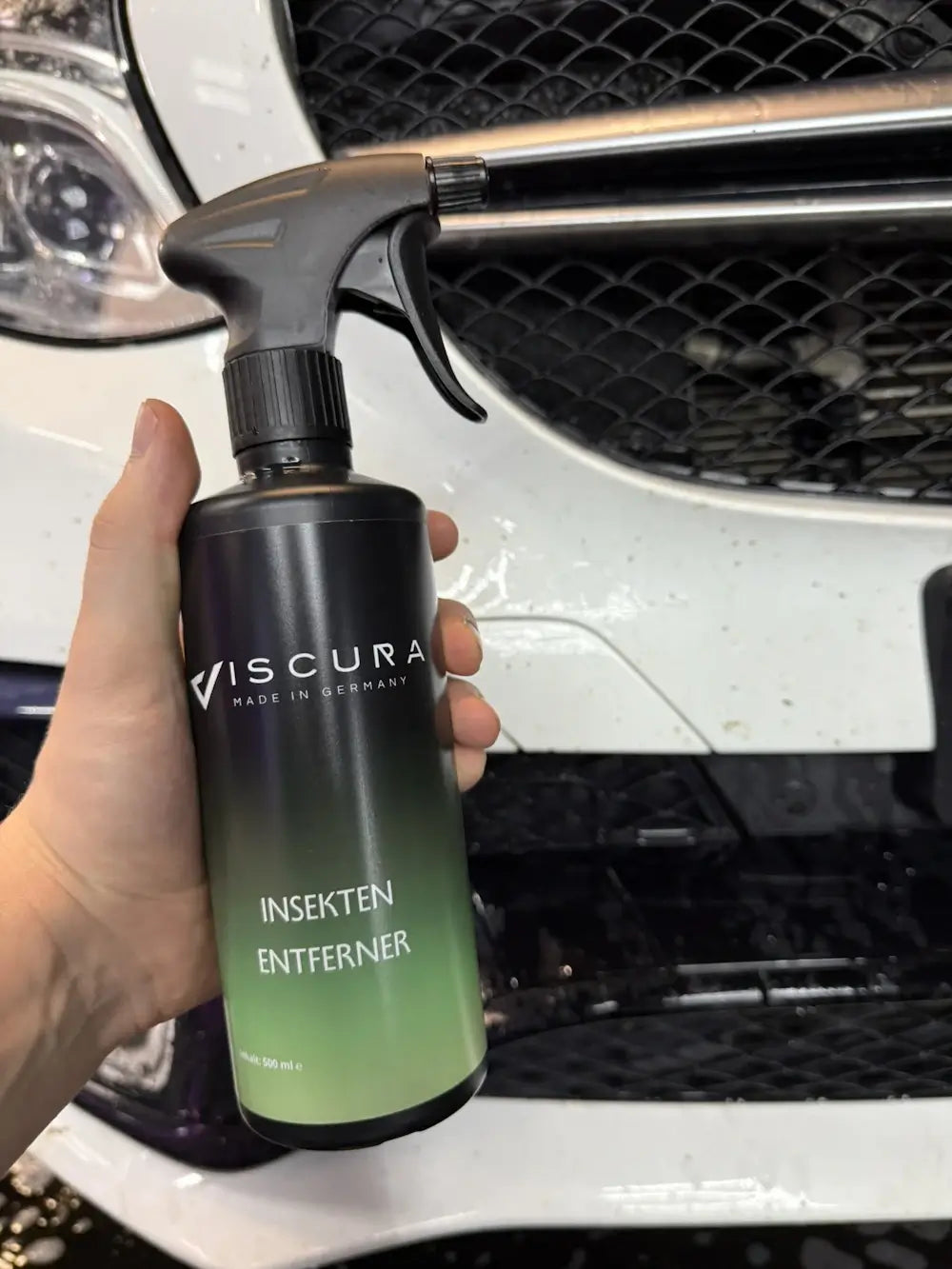 Hand holding VISCURA Insektenentferner spray bottle in front of a vehicle's grille.
