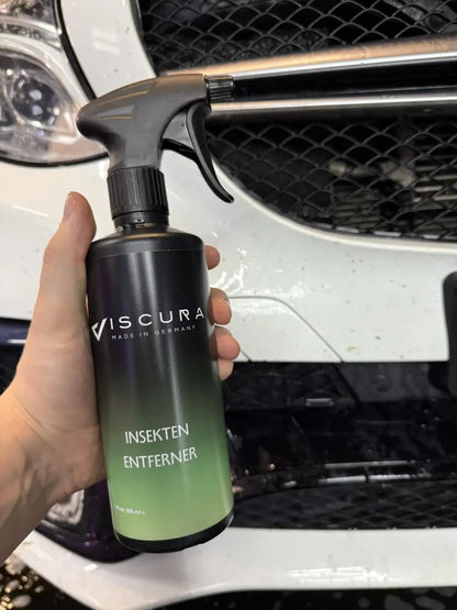 Hand holding VISCURA Insektenentferner spray bottle in front of a vehicle's grille.